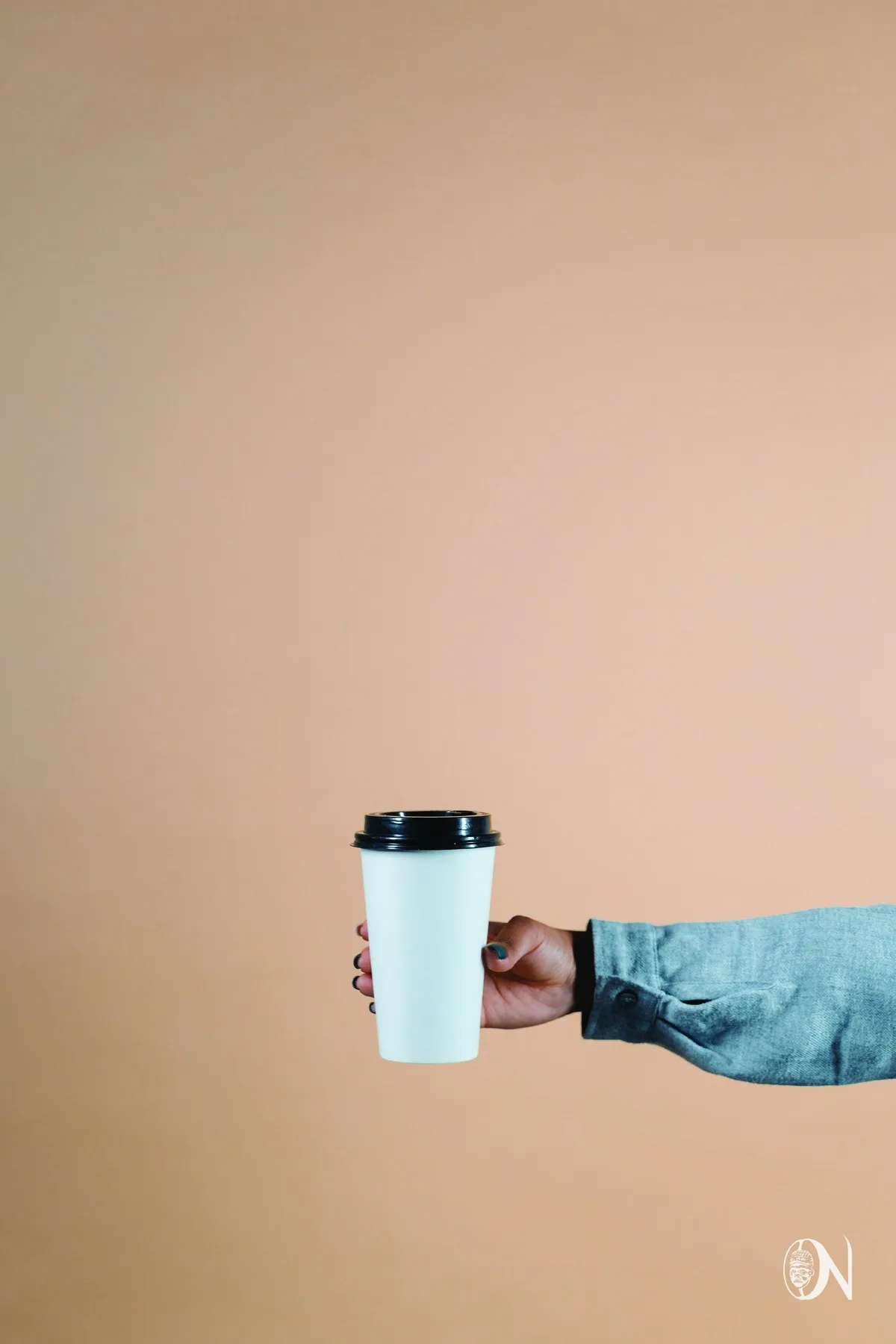 A hand holding an Okú Noir takeaway cup against a warm camel wall